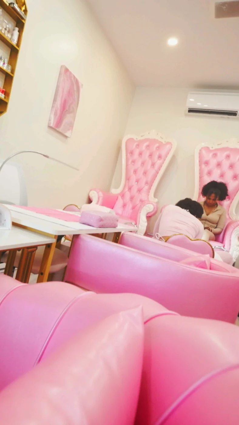 Tufted pink throne chairs seen low across glossy pink sofas, where a technician in pink works on a seated client’s feet