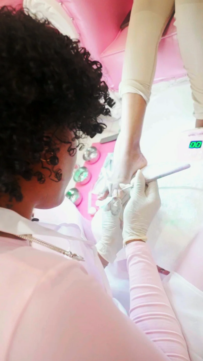 A gloved technician in pink, seen over the shoulder, files a client’s foot at a pink pedicure station in soft light